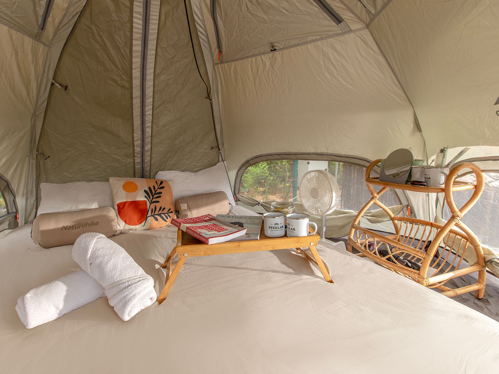 A cozy tent interior with a bed, pillows, a tray with books and mugs, a fan, and a wicker chair.