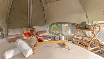 A cozy tent interior with a bed, pillows, a tray with books and mugs, a fan, and a wicker chair.