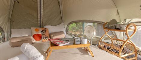 A cozy tent interior with a bed, pillows, a tray with books and mugs, a fan, and a wicker chair.