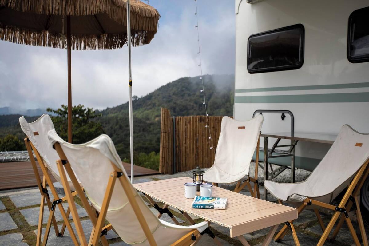 An outdoor seating area with a table and chairs, a thatched umbrella, and a view of a mountainous landscape.