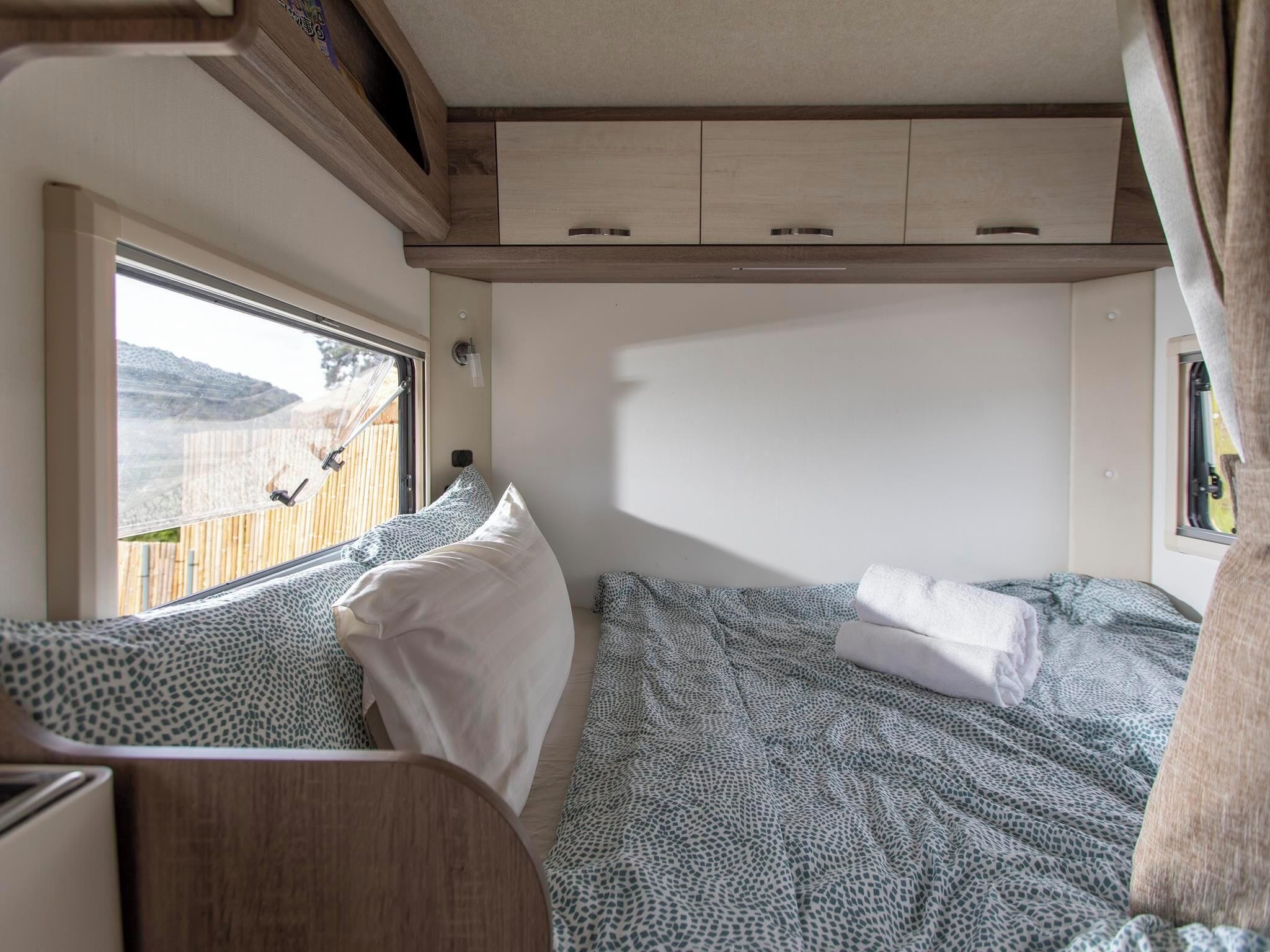 A compact RV bedroom with a bed, pillows, and a window overlooking a wooden deck.
