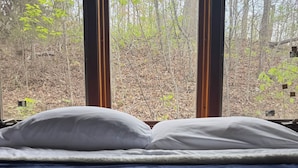 Ein Bett mit weißen Kissen und einer dunkelblauen Tagesdecke, mit Blick auf einen Wald durch ein Fenster.