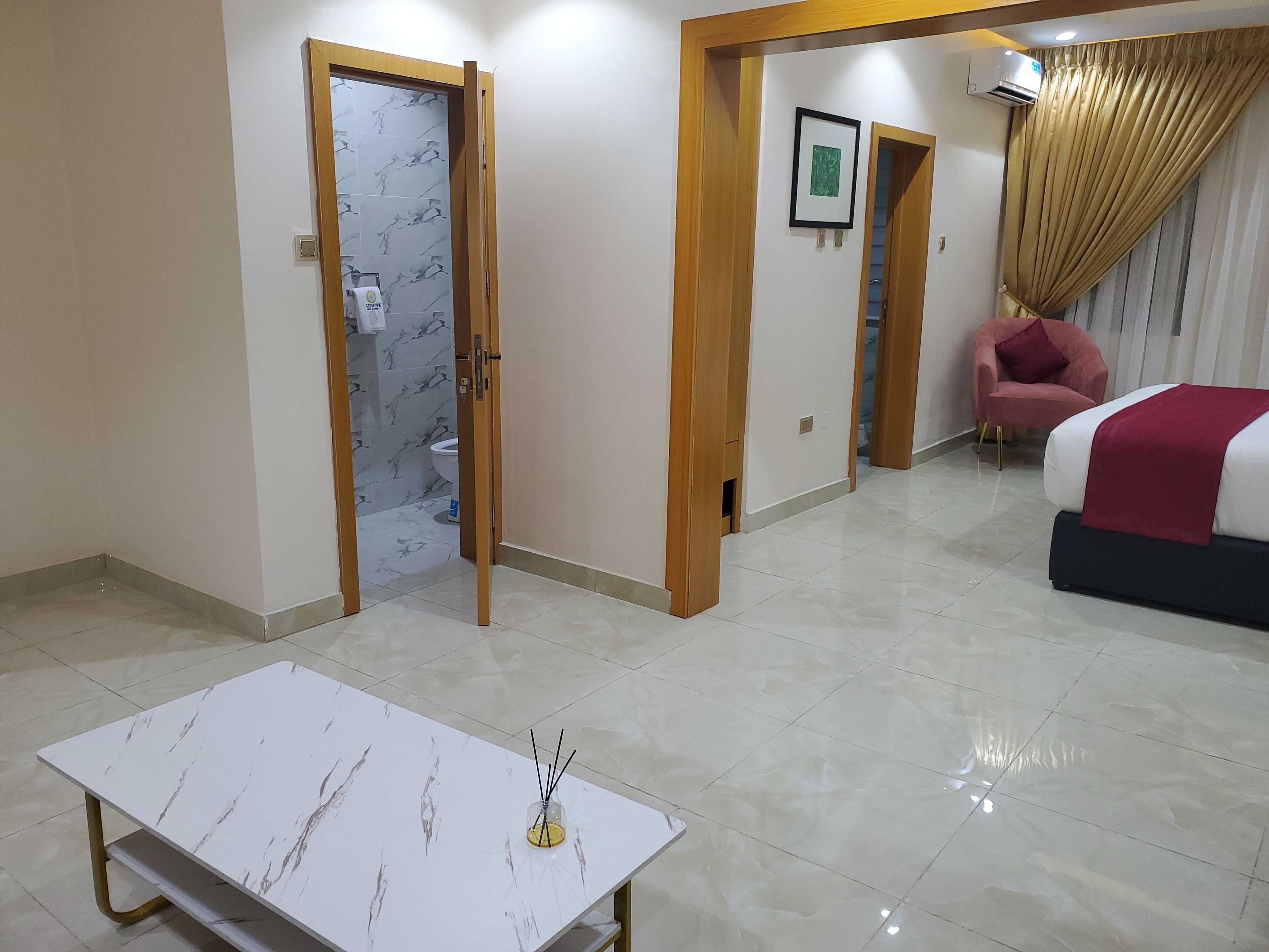 A hotel room with a marble-tiled floor, a bed with a red blanket, a pink chair, and a small table with a vase of flowers.