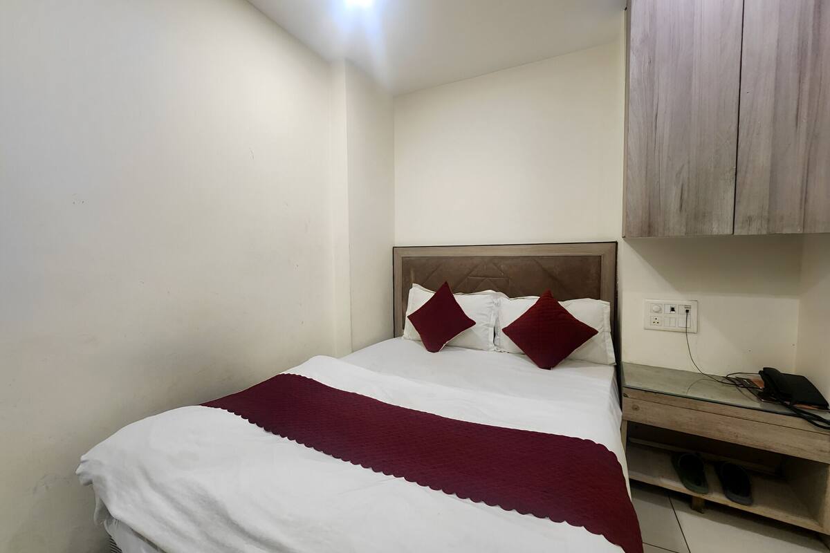 A small, neatly made bed with white and maroon bedding, a wooden headboard, and a bedside table with a phone and charger.