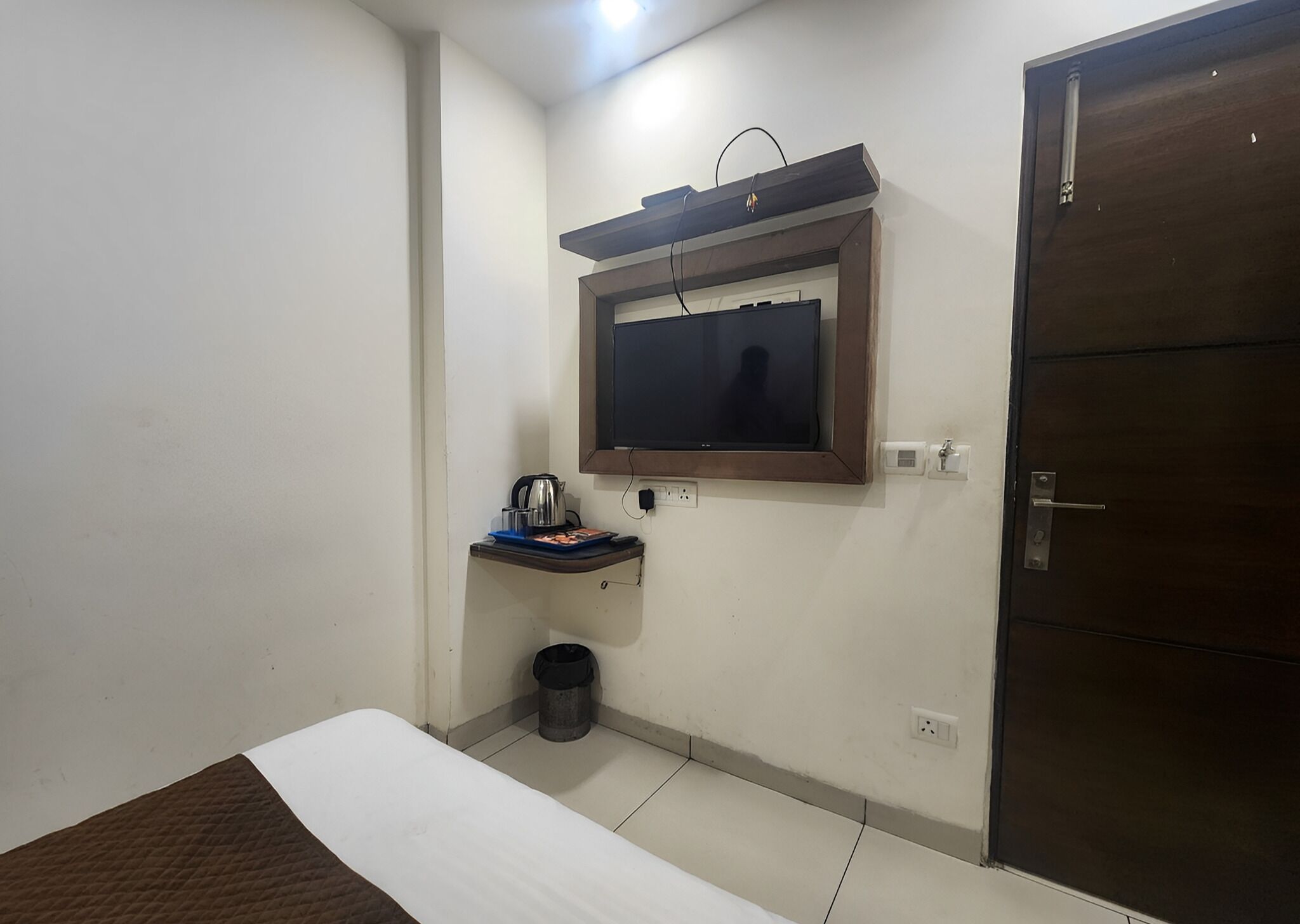 A small, simple room with a bed, a TV mounted on the wall, a small shelf with a kettle and a trash bin, and a door.