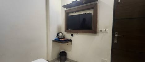 Ein kleiner, schlichter Raum mit einem Bett, einem an der Wand befestigten Fernseher, einem kleinen Regal mit einem Wasserkocher und einem Mülleimer sowie einer Tür.