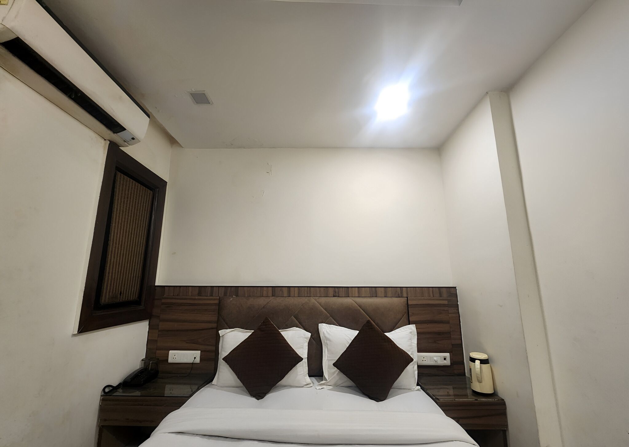 Une petite pièce propre, avec un lit simple, une table de chevet, une lampe de chevet et un radiateur fixé au mur.