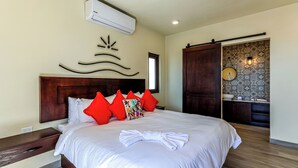 A hotel room with a large bed, a wooden headboard, a wall-mounted air conditioner, a bathroom with a sink and mirror, and a patterned tile backsplash.