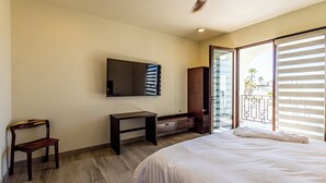 A bedroom with a bed, a wooden chair, a small table, a TV mounted on the wall, and a sliding glass door leading to a balcony.