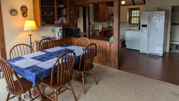 A cozy kitchen with wooden cabinets, a dining table with chairs, a refrigerator, and a microwave. - Pet Friendly Waterfront Overlooking Oyster River (Chatham)