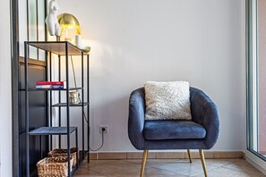 Ett modernt rum med en mörkblå fåtölj, en vit, fluffig kudde och en svart metallhylla med böcker och korgar.