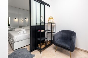 A modern bedroom with a bed, a black shelving unit with baskets, a dark armchair, and a bedside table with a lamp. - 49-"Le beach", Plein centre, climatisé, refait à n (Menton)