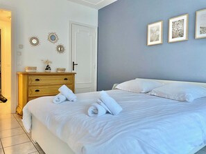 Un letto rifatto con lenzuola bianche e asciugamani arrotolati, un comò di legno con specchi decorativi e una porta con l'interruttore della luce.