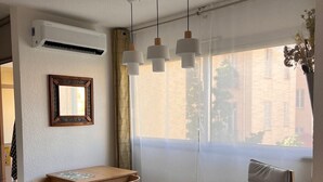 A modern living room with a wooden dining table, two chairs, a sofa, and a small side table with a magazine. There is a large window with a view of buildings outside. - 72-"Charmant studio" Plein centre, Calme, Terrasse (Menton)