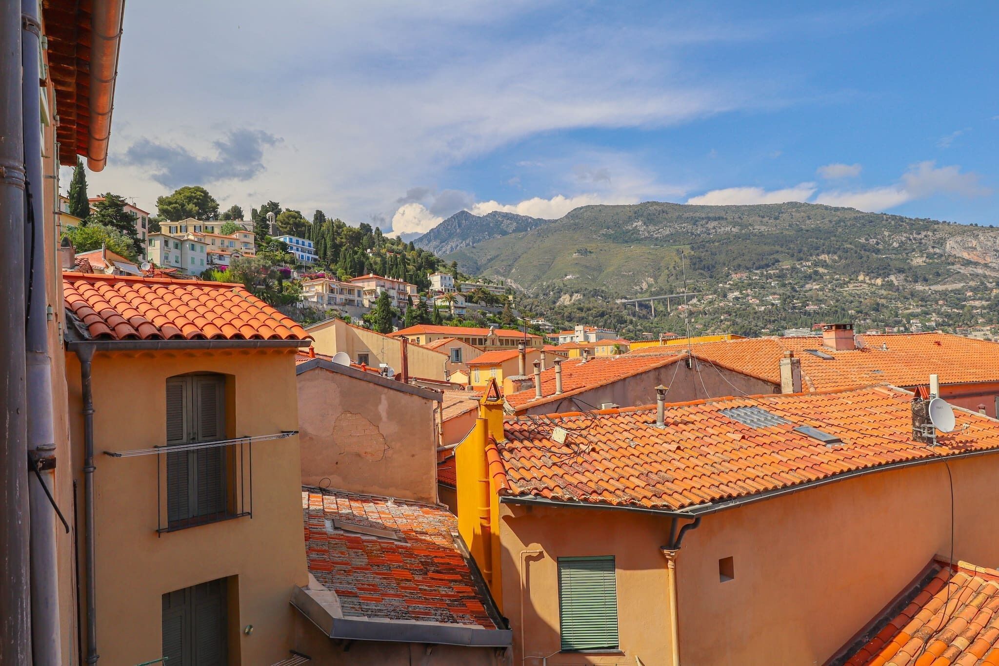 A residential area with terracotta-tiled roofs, a mountain in the background, and a clear sky.