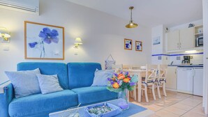 Un salon avec un canapé bleu, une table à manger avec des chaises, un coin cuisine et un climatiseur fixé au mur.