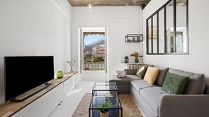 A modern living room with a flat-screen TV, a glass coffee table, a grey sofa, and a large window with a view of buildings and hills. - 82 -L’Écrin d’Alexandra, cosy 2 pièces (Menton)