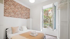 A bedroom with a brick wall, a bed with white bedding, a window with a view of greenery, and a white door. - 82 -L’Écrin d’Alexandra, cosy 2 pièces (Menton)