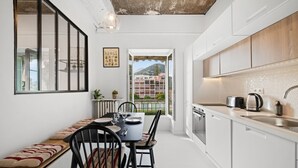 A modern kitchen with white cabinets, a dining area with a bench, and a view of a mountain through a window. - 82 -L’Écrin d’Alexandra, cosy 2 pièces (Menton)