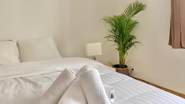 Ein ordentlich bezogenes Bett mit weißen Laken, ein Blumentopf, eine Lampe und ein gerahmtes Bild an der Wand.