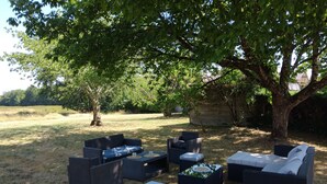 Un área de estar al aire libre con mobiliario de mimbre, incluyendo sofás y otomanas, bajo un árbol grande.