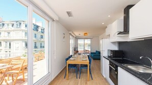 Une cuisine moderne avec un plan de travail noir et un évier en acier inoxydable, un coin repas avec une table en bois et des chaises bleues, et de grandes fenêtres offrant une vue sur un immeuble.
