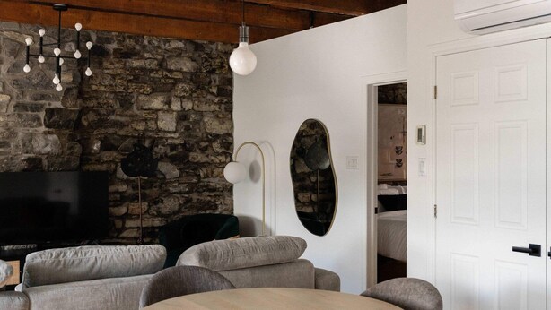 Una sala de estar moderna con pared de piedra, vigas de madera, una mesa redonda y sillones tapizados en gris.