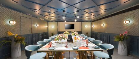 Un salón de conferencias con una larga mesa dispuesta para una reunión, rodeado de sillas y un diseño moderno del techo.