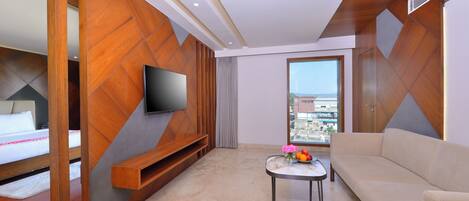Habitación de hotel con cama, televisor montado en la pared, sofá, mesita con un florero y un ventanal con vistas a la ciudad.