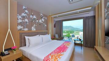 A hotel room with a large bed, a desk with a lamp, a chair, a small table with flowers, and a view of the city through a large window.
