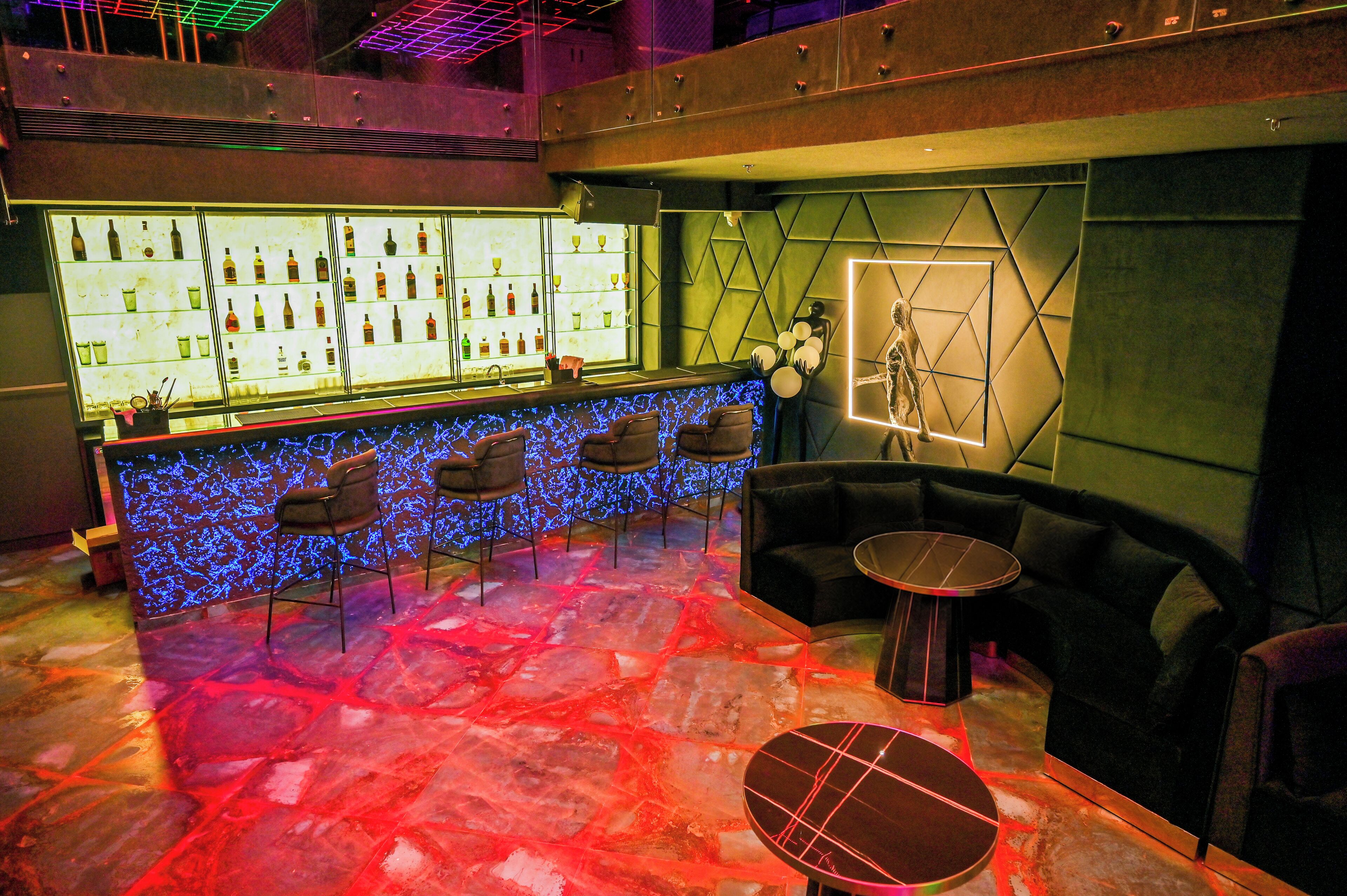A bar area with a counter displaying bottles and glasses, seating with a geometric wall design, and a floor with a red and blue light pattern.