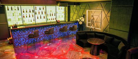 Zona de bar con mostrador que exhibe botellas y copas, asientos con diseño de pared geométrico y suelo con patrón de luces rojas y azules.