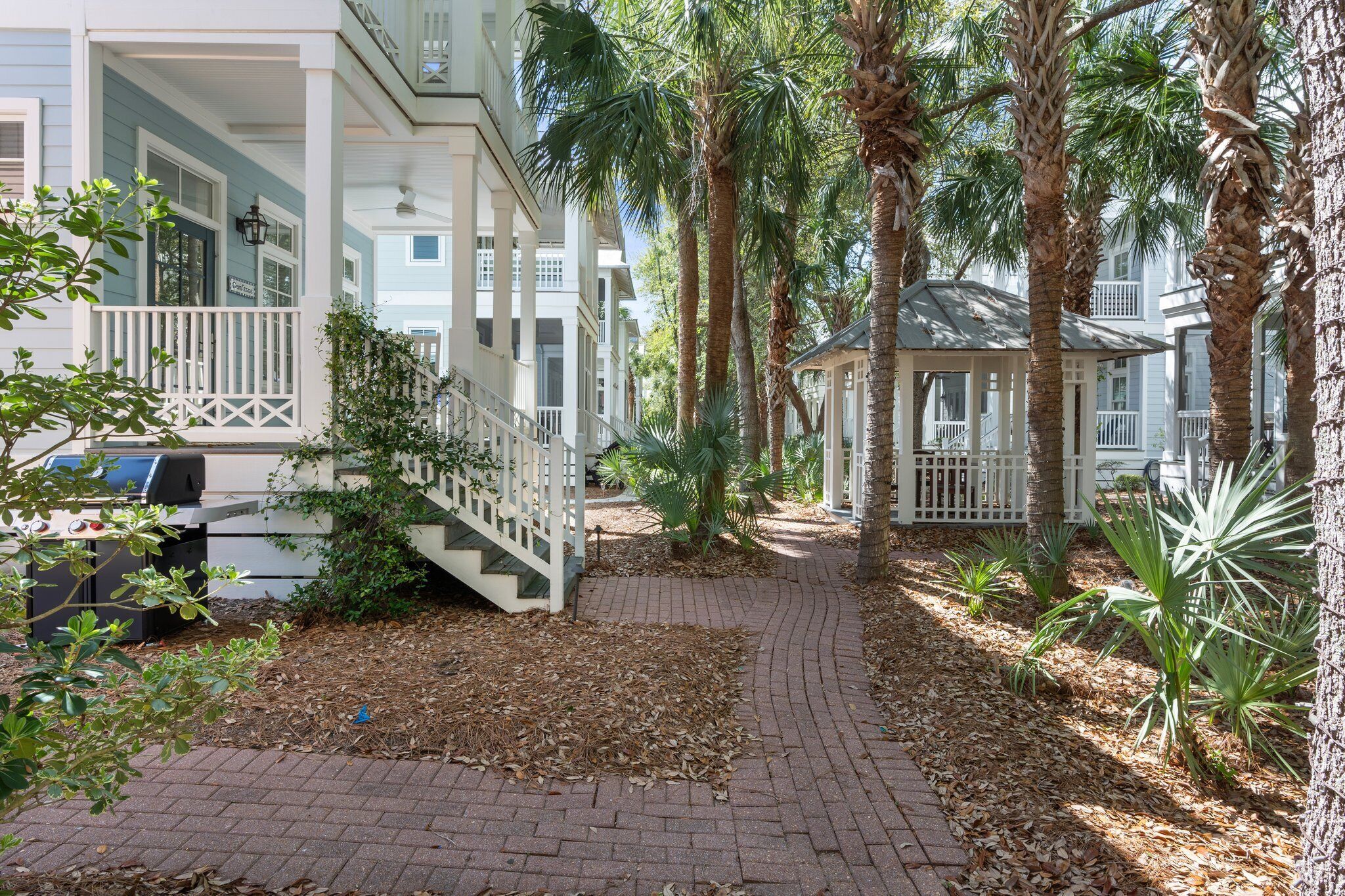 A pathway flanked by palm trees and a brick walkway leading to a building with a porch and a gazebo.