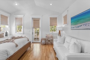 Un dormitorio con una cama, un sofá blanco, una silla de mimbre y un ventanal con vistas a la vegetación.