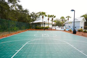 Una cancha de baloncesto exterior con superficie verde, rodeada por una valla y palmeras.