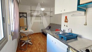 A compact kitchen with a dining area, a hanging light fixture, and a small window with curtains. - Studio 2 personnes en rdc quartier de la Corniche (Sète)