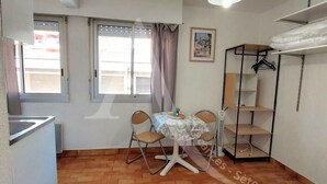 A compact room with a dining table, chairs, a small shelf unit, and a window with curtains. - Studio 2 personnes en rdc quartier de la Corniche (Sète)