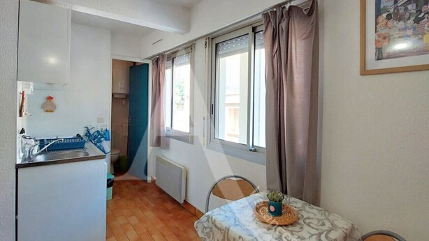 A compact kitchen with a refrigerator, sink, and blue dish rack. A dining table with a patterned tablecloth and a small potted plant is set against a window with curtains. - Studio 2 personnes en rdc quartier de la Corniche (Sète)