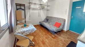 A compact living space with a sofa, a small table, and a hanging shelf. - Studio 2 personnes en rdc quartier de la Corniche (Sète)