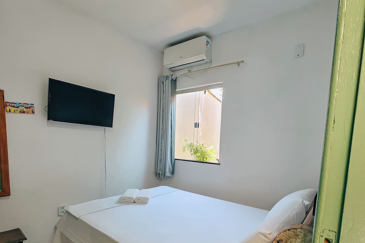 A clean, well-lit hotel room with a bed, a TV, an air conditioning unit, and a window with curtains.