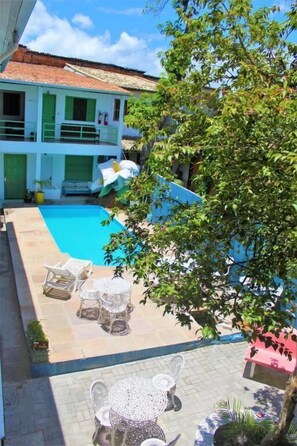 A pool area with lounge chairs and a table, surrounded by a building with a tiled roof and a tree. - Pousada Rio do Prado (Porto Seguro)