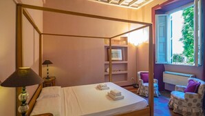 A four-poster bed with a white bedspread, two bedside tables with lamps, a built-in shelf with books, a window with an open view, and two armchairs with purple cushions. - CAMPO DE' FIORI PINKY HOUSE (Rome)