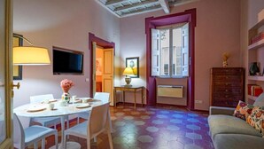 A cozy room with a dining table, chairs, a sofa, a TV, a window with shutters, and a patterned floor. - CAMPO DE' FIORI PINKY HOUSE (Rome)