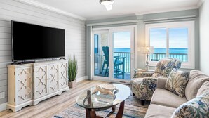 A living room with a flat-screen TV, a glass coffee table, a sofa, and a view of the ocean through large windows. - 723- Beautiful, TOP FLOOR UNIT w/ Stunning Views! Free Beach Service (Destin)