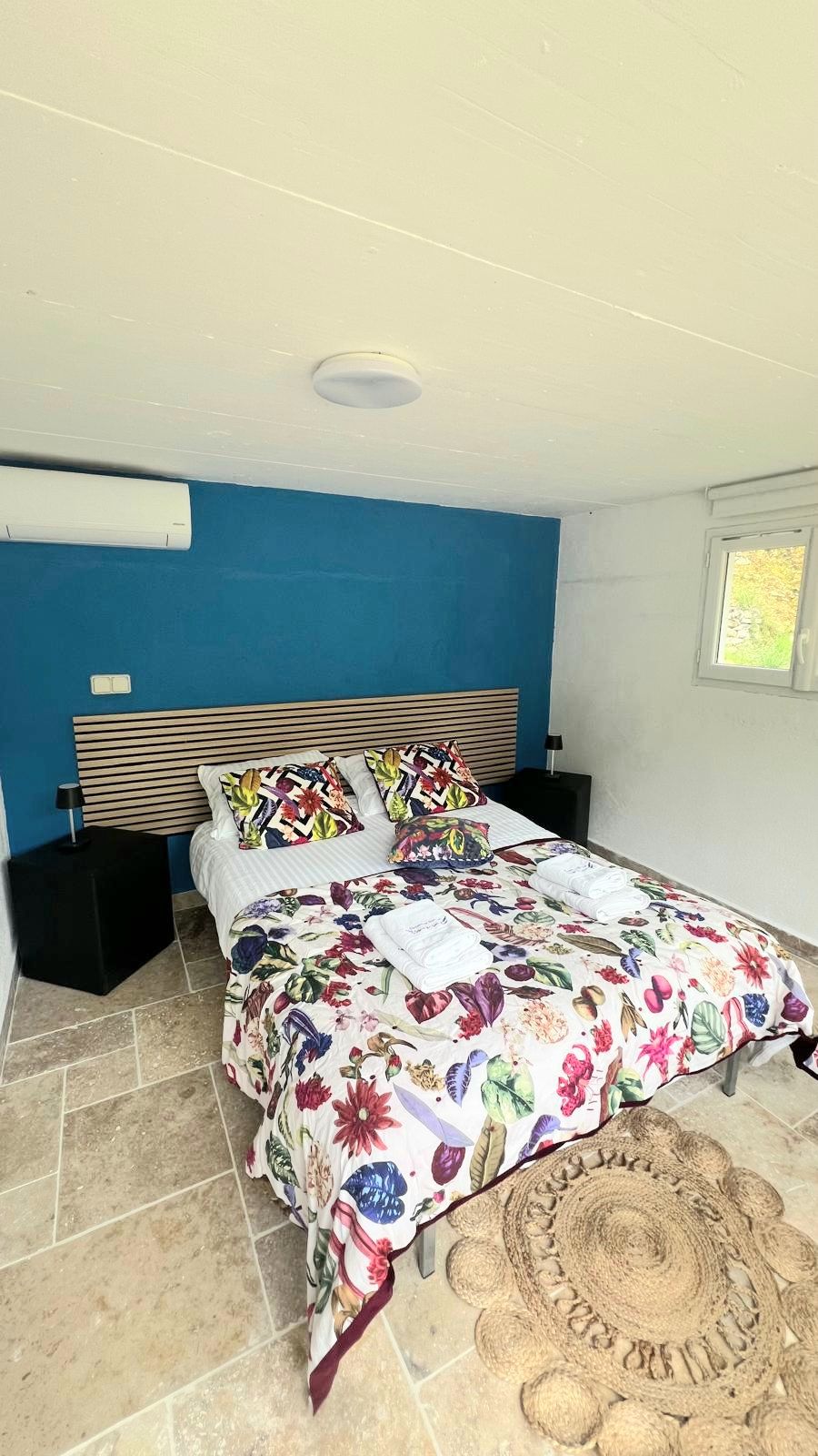 A bedroom with a bed, a headboard, a nightstand, a window, and a ceiling vent.