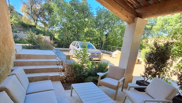 Een overdekt terras met een bank, stoelen en een tafel, met uitzicht op een tuin en een geparkeerde auto.