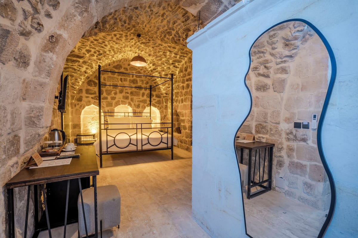 A stone-walled room with a bed, a table with a lamp, and a mirror.