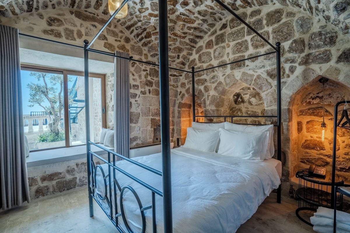 A stone-walled bedroom with a four-poster bed, a view through a window, and a stone fireplace.