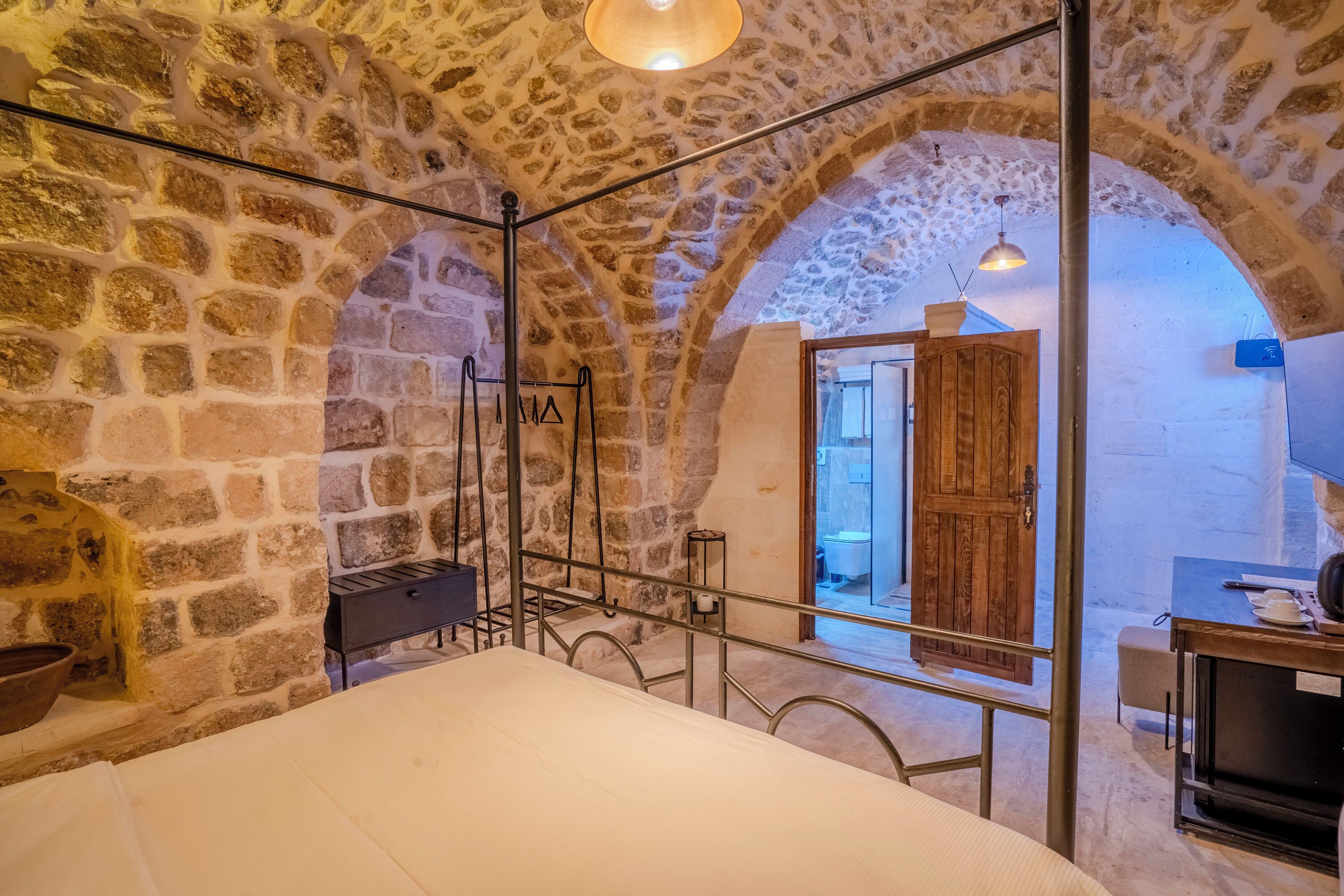 A rustic stone-walled room with a bed, a wooden door, and a small table with a cup and saucer.