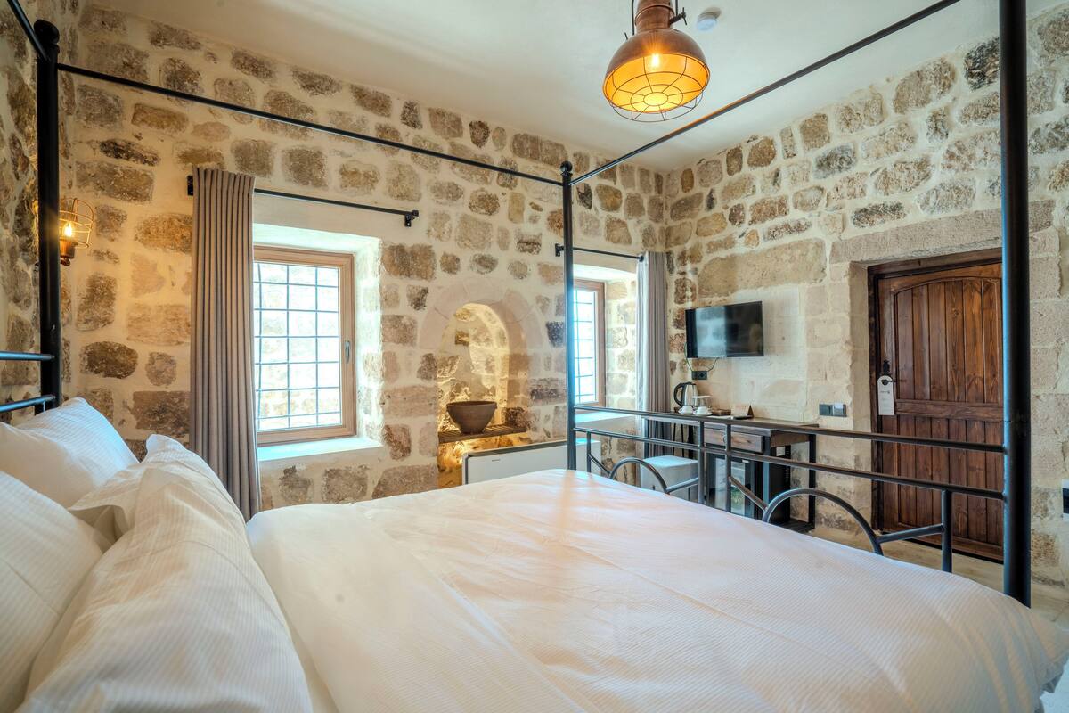 A bedroom with a stone wall, a bed, a desk, and a television.